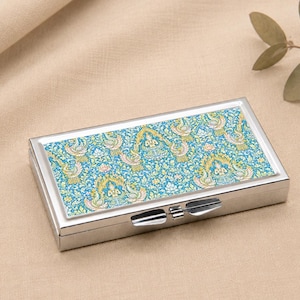 May include: A rectangular silver metal case featuring a hinged lid with a vibrant blue and gold patterned inlay. The design displays intricate repeating motifs of stylised figures and floral elements across the top surface of the metallic container.