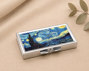 Puede incluir: Un pastillero rectangular plateado con una impresión de "La noche estrellada" de Van Gogh. La obra de arte representa un cielo nocturno arremolinado con estrellas amarillas y un pueblo oscuro. La caja tiene una tapa con bisagras y un cierre.