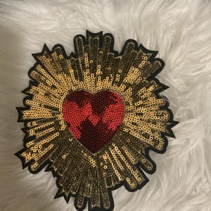 May include: A decorative patch featuring a central red sequined heart surrounded by radiating gold sequined rays. The entire design is outlined in black, creating a sharp contrast against the surrounding texture of the embroidered sunburst pattern.