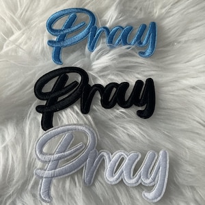 May include: Three embroidered patches featuring the word Pray in a cursive script font. The top patch is blue, the middle patch is black, and the bottom patch is white. Each patch has a raised, stitched texture with a distinct border outline.