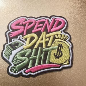 Könnte beinhalten: Ein bestickter Aufnäher mit dem Text SPEND DAT SHIT in rosa und gelbem Garn. Das Design enthält einen grünen Stapel Geldscheine auf der linken Seite und einen gelben Geldsack mit schwarzem Dollarzeichen rechts, alles schwarz-weiß umrandet.