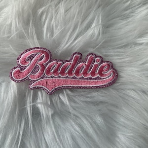 May include: An embroidered patch featuring the word Baddie in a cursive script font. The lettering is pink with a glittery texture, outlined in a darker pink thread, and set against a matching pink glittery background with a swoosh underline.