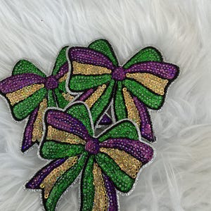 May include: Three decorative bow-shaped patches covered in shimmering sequins. Each bow features a striped pattern of vibrant green, deep purple, and metallic gold sequins, with a silver sequined border outlining the entire shape of the bows.