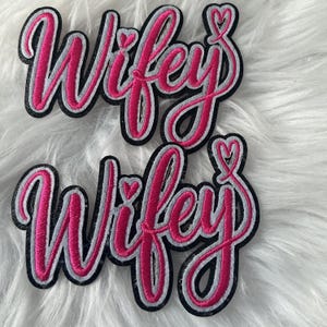 May include: Two embroidered patches featuring the word Wifey in a bright pink cursive script. Each patch has a white outline and a thick black border, with a small heart icon integrated into the letter y at the end of the word.