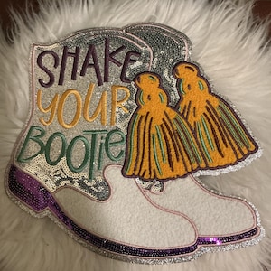 Op de afbeelding: Een decoratieve patch in de vorm van een cowboylaars, bedekt met zilveren lovertjes met een paarse rand. De tekst SHAKE YOUR BOOTIE is geborduurd in paars, goud en groen. Twee goudkleurige kwastjes zijn bevestigd aan de bovenkant van de laars.