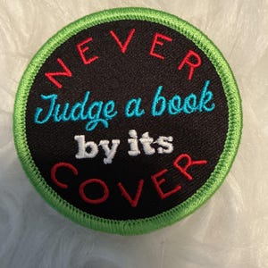 Puede incluir: Un parche bordado circular con fondo negro y borde verde brillante. El texto "NEVER" y "COVER" está bordado en rojo, "Judge a book" en azul claro y "by its" en blanco, todo centrado en la superficie del parche.