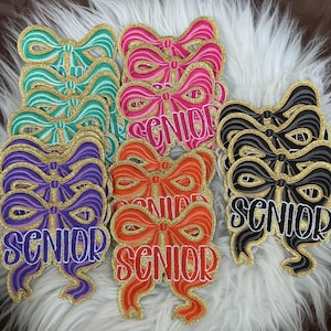 Op de afbeelding: Een verzameling geborduurde strikvormige patches in de kleuren groenblauw, roze, paars, oranje en zwart. Elke patch heeft een gouden glitterrand en het woord SENIOR in witte letters in het midden van het strikontwerp.