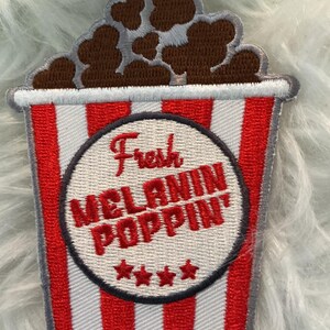 May include: Embroidered patch of a popcorn bucket design. The bucket has red and white stripes, with a central circle reading "Fresh MELANIN POPPIN'" in red. Brown popcorn is at the top, and four red stars are below the text.
