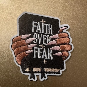 May include: An embroidered patch featuring a black book with the white text FAITH OVER FEAR and small cross symbols. Two brown hands with long, light-coloured pointed fingernails are wrapped around the book, which has two black bookmarks hanging down.