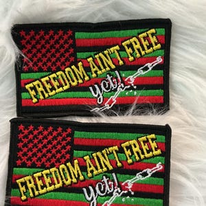 Op de afbeelding: Twee rechthoekige patches met een zwarte rand. Elke patch heeft een rood-zwart vlagontwerp met de tekst "FREEDOM AIN'T FREE yet!" in geel en wit. Er is ook een afbeelding van een gebroken ketting.