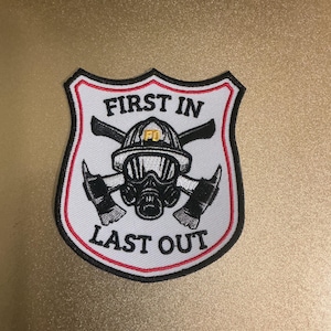 May include: A shield-shaped embroidered patch with a white background and black and red borders. It features a central graphic of a firefighter helmet and gas mask crossed with two axes. The text FIRST IN and LAST OUT is stitched in black.