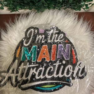 May include: A large, black, sequined patch featuring the text "I'm the MAIN Attraction" in multi-colored, embroidered lettering. The word "MAIN" is highlighted in teal, orange, and purple sequins, while the remaining text is in white.