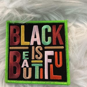 May include: Embroidered patch with the words "BLACK IS BEAUTIFUL" in colorful letters on a black background. The patch has a green border and is likely for clothing or accessories.