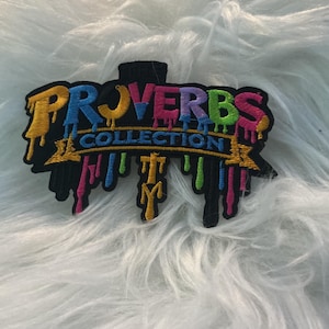 Puede incluir: Un parche bordado negro con el texto "THE PROVERBS COLLECTION" en un estilo de pintura que gotea multicolor. Las letras están rellenas con hilo amarillo, azul, rosa y verde, con gotas verticales adicionales que se extienden desde la parte inferior.