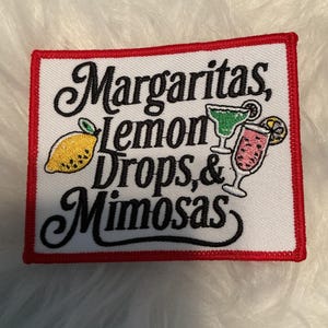 May include: A rectangular embroidered patch with a white background and a red border. The text reads Margaritas, lemon Drops, & Mimosas in black script. It features a yellow lemon, a green cocktail in a glass, and a pink drink with a citrus slice.