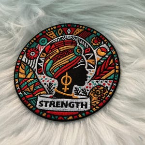 May include: Embroidered patch featuring a profile of a woman with a head wrap, surrounded by colorful geometric designs. The patch includes the word "STRENGTH" in a white rectangular banner. The patch is circular with a black border.