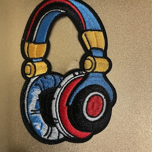 May include: An embroidered patch shaped like a pair of over-ear headphones. The design features a blue headband with yellow accents, and ear cups colored in blue, white, red, and black, all outlined with a thick black border for definition.