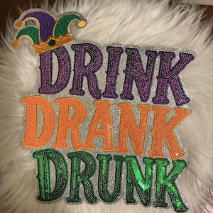 May include: A decorative patch featuring the text DRINK DRANK DRUNK in purple, orange, and green sequins on a silver glitter background. A jester hat icon in purple, green, and gold sits above the text. The design is outlined in silver glitter.