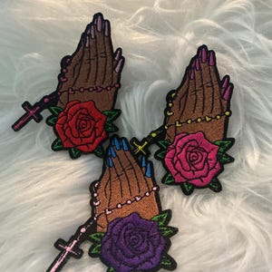 May include: Three embroidered patches featuring brown hands in a praying position. Each patch includes a rosary with a cross and a flower at the base. One patch has a red rose, one a pink rose, and one a purple rose with green leaves.