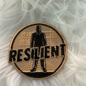 Op de afbeelding: Een cirkelvormige patch met een gouden rand en een bruinachtig midden. De patch toont een zwart silhouet van een persoon die staat, met het woord "RESILIENT" in zwart over het midden. De cijfers "1920" staan boven en onder.