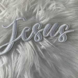Op de afbeelding: Een witte geborduurde patch met het woord Jesus in een cursief lettertype. De letters hebben een verhoogde textuur met een duidelijke gestikte rand, wat zorgt voor een strakke en gedefinieerde uitstraling van de tekst.