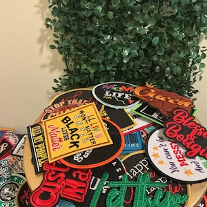 May include: A collection of assorted embroidered fabric patches featuring various shapes, colors, and text designs. The patches include phrases such as "Black Lives Matter," "Mom Life," "Queen," "Blessed and Boujee," and "Let them," with diverse fonts and graphic elements.