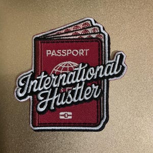 Puede incluir: Un parche bordado rectangular que muestra un pasaporte de color rojo oscuro con un icono de globo terráqueo blanco y el texto PASSPORT. Superpuesto en letra cursiva negra con borde blanco aparece el texto International Hustler. El diseño tiene borde.
