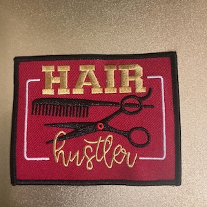 Puede incluir: Un parche rectangular bordado con fondo rojo y borde negro. El texto HAIR está bordado en dorado y la palabra hustler en letra cursiva dorada. Un peine negro y unas tijeras negras están bordados en el centro, enmarcados por una línea blanca.