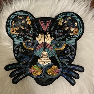 Può includere: Una toppa decorativa a forma di testa di felino, realizzata in tessuto nero e decorata con paillettes e ricami iridescenti, dorati, rosa e turchesi. Il design presenta cuciture dettagliate per gli occhi, il naso e i baffi.