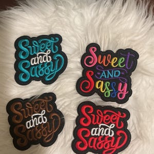 May include: Four embroidered patches with black backgrounds, each featuring the text "Sweet and Sassy" in different color schemes. One patch has teal and white thread, one has multi-colored rainbow thread, one has brown and white, and one has red and white.
