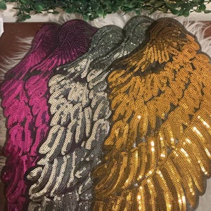 Peut inclure: Un ensemble de quatre écussons en forme d'ailes recouverts de paillettes scintillantes. Les écussons sont disposés en éventail et présentent des couleurs distinctes, dont le magenta vif, l'argenté métallique, le gris anthracite et l'or brillant.