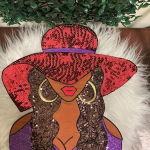 May include: A decorative applique featuring a stylized figure wearing a wide-brimmed red sequin hat and a purple sequin top. The figure has brown skin, long dark brown wavy hair, bright red lips, and large gold hoop earrings, all rendered in fabric.
