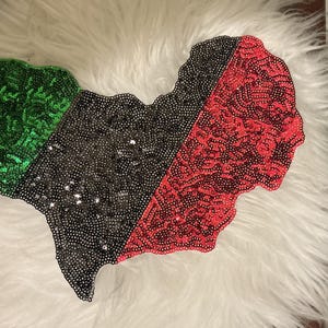 Peut inclure: Un écusson décoratif en forme de carte du Nigeria, présentant un design coloré avec des sections verte, noire et rouge. Toute la surface est densément recouverte de petites paillettes réfléchissantes qui créent une finition texturée.