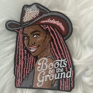 May include: An embroidered patch featuring a person with long pink braided hair wearing a dark red cowboy hat with silver accents. The patch includes the white embroidered text "Boots on the Ground" and has a black border outline.