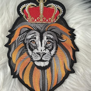 May include: An embroidered patch featuring a lion head with a thick mane in shades of orange, gray, and black. The lion is wearing a red and gold crown with white bead-like accents. The entire design is outlined in black thread on a fabric base.
