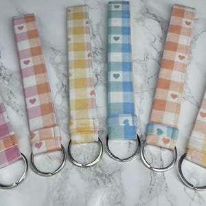 May include: Six fabric keychains in pastel colors: pink, peach, yellow, and blue. Each keychain features a gingham pattern with small heart accents and a silver metal ring. The keychains are arranged side by side.