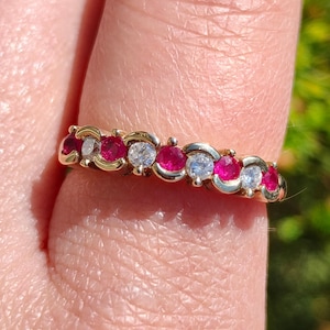 May include: A gold band ring with alternating red rubies and white diamonds. The ring is set in a delicate, curved design.