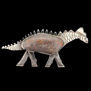 May include: A dinosaur-shaped brooch crafted from polished silver metal, featuring a large, textured, oval-shaped stone inlay in shades of brown, grey, and orange. The body has a row of triangular spikes along the back and detailed etched lines.