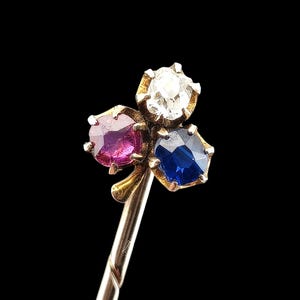 May include: A gold pin with three gemstones: a clear diamond, a deep blue sapphire, and a magenta ruby. The gemstones are set in a floral design. The pin has a long, slender stem, against a black background.