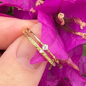 May include: A gold ring with a small diamond is held against a backdrop of vibrant purple bougainvillea flowers. The ring has a delicate design with a small diamond and a dotted band. The flowers are a bright magenta color with green leaves.
