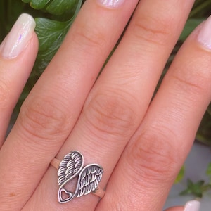 Peut inclure: Bague argent&eacute;e repr&eacute;sentant des ailes d'ange. Les ailes sont orn&eacute;es de d&eacute;tails de plumes et un petit c&oelig;ur est visible &agrave; la base. La bague est pr&eacute;sent&eacute;e sur un doigt, sur fond vert.