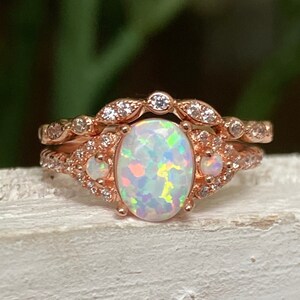 May include: A rose gold ring set featuring a central oval-cut opal with iridescent flashes of color, flanked by two smaller round opals. The bands are adorned with numerous small, clear pavé-set stones arranged in a delicate, intricate pattern.
