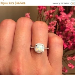 May include: A silver ring with a square-shaped opal gemstone surrounded by a halo of smaller diamonds. The ring is on a person's finger.