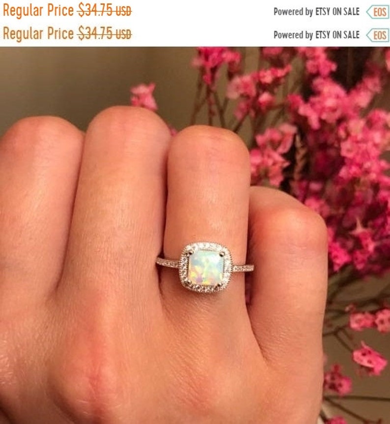 May include: A silver ring with a square-shaped opal gemstone surrounded by a halo of smaller diamonds. The ring is on a person's finger.