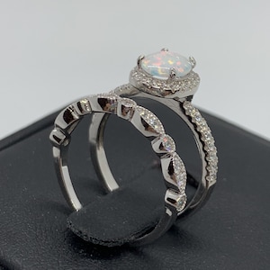 May include: A silver ring set with a white opal center stone surrounded by smaller clear stones. The engagement ring has a halo setting and the wedding band has a delicate, scalloped design.