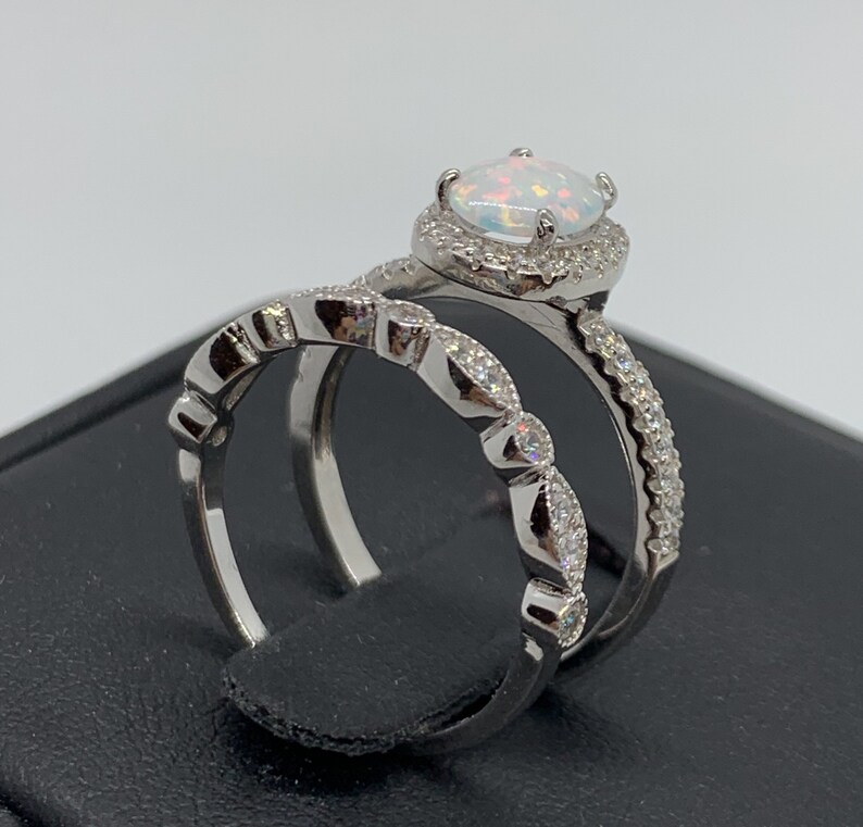 May include: A silver ring set with a white opal center stone surrounded by smaller clear stones. The engagement ring has a halo setting and the wedding band has a delicate, scalloped design.