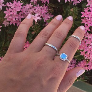 May include: A hand wearing three silver rings. The ring on the middle finger has a square blue opal stone with a halo of smaller stones. The other two rings are thin and have a beaded design.