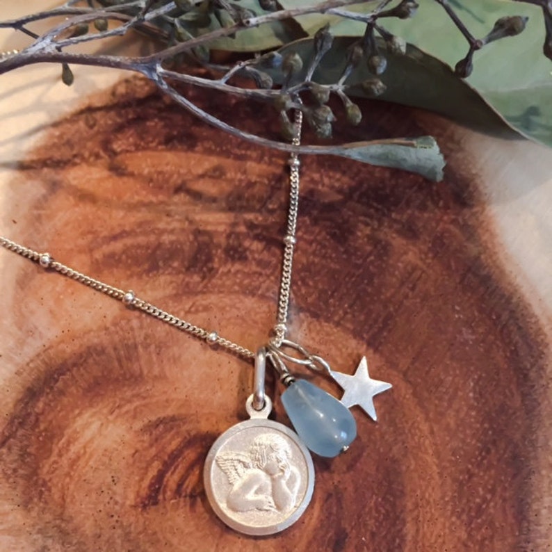 May include: A silver necklace with a charm of an angel, a blue gemstone, and a star. The necklace is on a wooden surface.