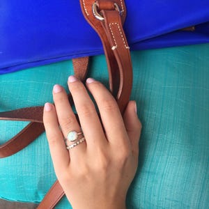 May include: A hand wearing two rings, one with a white opal stone and the other with a silver band. The hand is resting on a turquoise fabric surface.