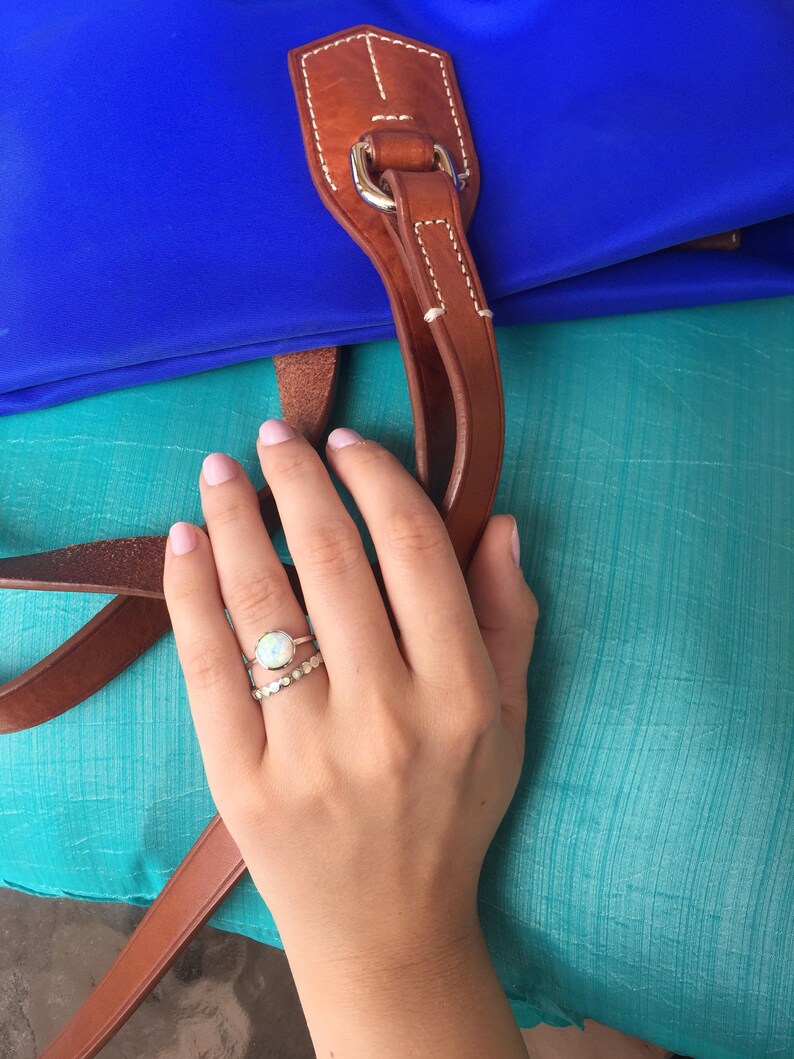 May include: A hand wearing two rings, one with a white opal stone and the other with a silver band. The hand is resting on a turquoise fabric surface.